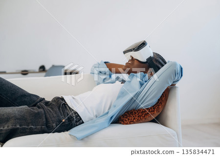 Resting on sofa with VR headset on. Woman is at home in domestic room 135834471