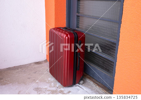 Red rolling suitcase stands next to apartment door. Luggage ready for travel or moving. Departure and arrival concept for journey. 135834725