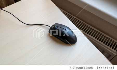Black wired computer mouse on a light wood desk, ready for use in office work or gaming. Technology device for home or professional setting. Black wired computer mouse on a light wood desk, ready for use in office work or gaming. Technology device for home or professional setting. 135834731