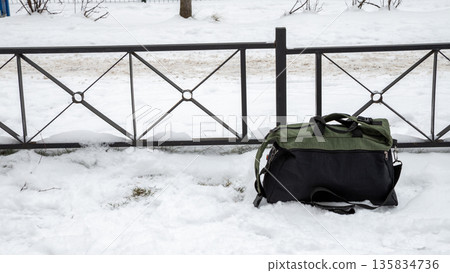 Travel bag abandoned in snow next to a metal fence. Lost luggage concept in winter environment. Traveling and journey theme. 135834736