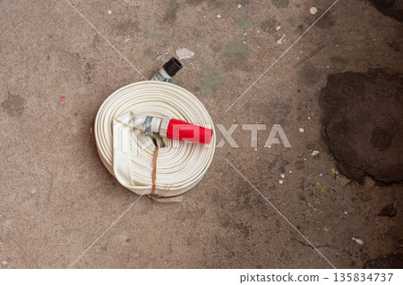 Rolled fire hose with a red nozzle on a concrete floor. Emergency equipment for fire safety and industrial protection. Firefighter tool for independent fire extinguishing Rolled fire hose with a red nozzle on a concrete floor. Emergency equipment for fire safety and industrial protection. Firefighter tool for independent fire extinguishing 135834737