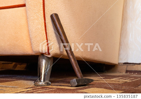 Hammer leaning against a beige sofa, next to a metal leg on a patterned carpet floor. A common household repair tool for various home maintenance tasks. 135835187
