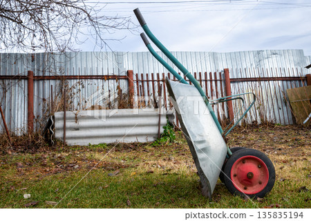 Empty garden wheelbarrow standing on grass next to a metal fence. outdoor work, gardening, or construction projects. profile view Empty garden wheelbarrow standing on grass next to a metal fence. outdoor work, gardening, or construction projects. profile view 135835194