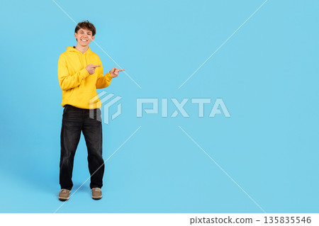 Smiling young man in yellow hoodie pointing sideways on blue studio background 135835546
