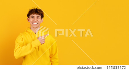 Happy guy in yellow hoodie pointing sideways at copy space, studio background 135835572