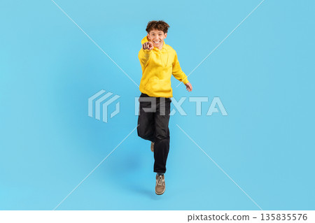 Smiling young man pointing directly at camera while jumping on blue background Smiling young man pointing directly at camera while jumping on blue background 135835576