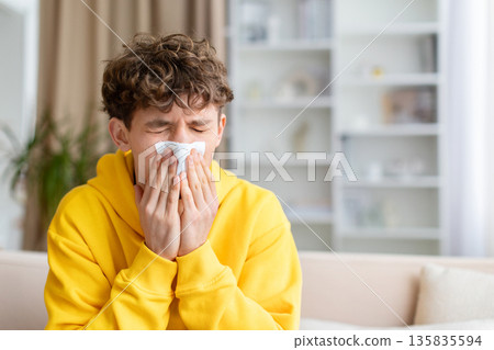 Sick teenage boy sneezing into tissue, sitting on couch at home 135835594