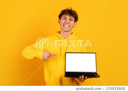 Smiling young man pointing at laptop with blank screen on yellow background Smiling young man pointing at laptop with blank screen on yellow background 135835608