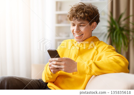 Smiling young man relaxing on sofa while texting on smartphone at home 135835618