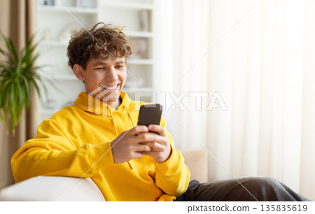 Smiling young man relaxing on sofa and using smartphone at home Smiling young man relaxing on sofa and using smartphone at home 135835619