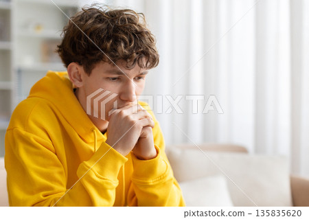 Thoughtful teenage boy sitting on sofa with hands near face Thoughtful teenage boy sitting on sofa with hands near face 135835620