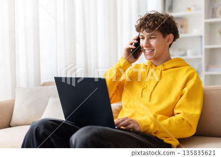 Teenage boy talking on smartphone while using laptop on couch at home Teenage boy talking on smartphone while using laptop on couch at home 135835621