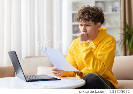 Thoughtful young man reading document while sitting with laptop at home Thoughtful young man reading document while sitting with laptop at home 135835622