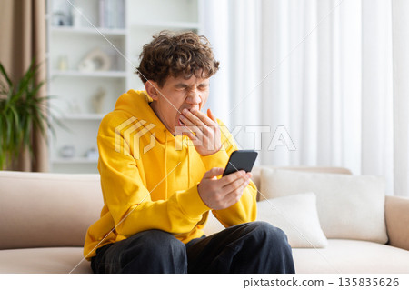 Tired teenage guy yawning while looking at smartphone screen at home 135835626
