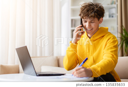 Cheerful young man talking on phone and writing notes at home desk Cheerful young man talking on phone and writing notes at home desk 135835633