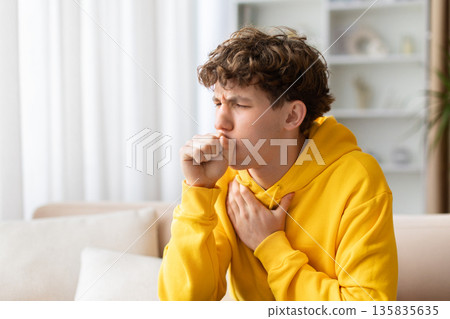 Teenage boy coughing with hand on chest, sitting on sofa at home Teenage boy coughing with hand on chest, sitting on sofa at home 135835635