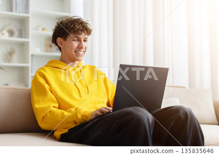 Smiling teen guy using laptop on sofa at home, enjoying online activity, Smiling teen guy using laptop on sofa at home, enjoying online activity, 135835646