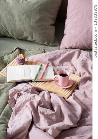 Open notebook with pens, burning candle, coffee cup on wooden tray in bed. 135835979