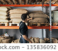 Back view of male warehouse worker standing at shelves and adjusting bags with coffee 135836092