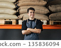 Portrait of a young male in apron with crossed hands against shelves. Cheerful employee looking away while standing at warehouse. 135836094