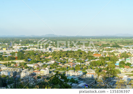 High angle view of a small Thai city with colorful rooftops, a traditional Buddhist temple, and a tall communication tower. Scenic suburban landscape with green trees and mountains under a blue sky. High angle view of a small Thai city with colorful rooftops, a traditional Buddhist temple, and a tall communication tower. Scenic suburban landscape with green trees and mountains under a blue sky. 135836397
