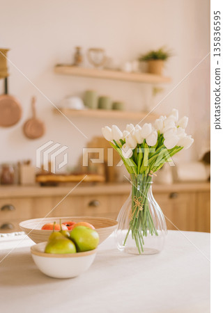 White tulips in a glass vase and fresh fruits on a table in a bright, cozy rustic kitchen interior. 135836595