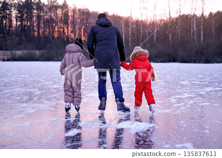 Father holding hands with two children while skating on frozen lake at winter sunset. Family outdoor activity, bonding, care, safety and togetherness in cold season nature landscape. Father holding hands with two children while skating on frozen lake at winter sunset. Family outdoor activity, bonding, care, safety and togetherness in cold season nature landscape. 135837611