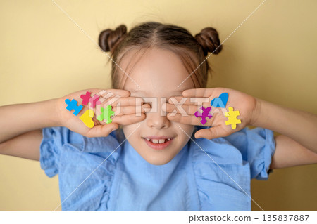 Smiling child covering eyes with colorful puzzle pieces and hearts. Autism awareness and inclusion concept for World Autism Awareness Day, education, support, diversity and social campaigns. Smiling child covering eyes with colorful puzzle pieces and hearts. Autism awareness and inclusion concept for World Autism Awareness Day, education, support, diversity and social campaigns. 135837887