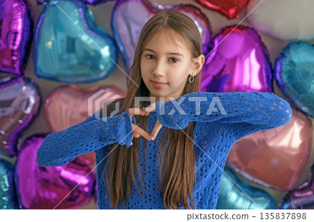Smiling child making heart shape with hands against colorful heart balloons. Valentines Day concept, love, kindness, childhood, affection, celebration and positive emotions. 135837898