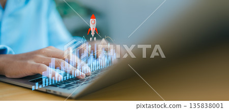 Person using laptop with business growth chart and rocket icon overlay. Concept of startup success, financial progress, market acceleration, and skyrocketing corporate achievement. 135838001