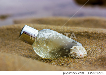 A small, clear, silver-capped cosmetic bottle and a seashell rest on the sand. Bubbles inside the bottle suggest a natural, beach-themed product for skincare. Serum, shampoo, shower gel on a sea coast A small, clear, silver-capped cosmetic bottle and a seashell rest on the sand. Bubbles inside the bottle suggest a natural, beach-themed product for skincare. Serum, shampoo, shower gel on a sea coast 135839276