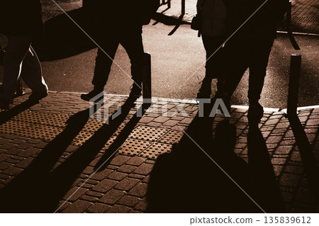 Candid, high-contrast shot of people's silhouettes and long shadows walking on a paved urban sidewalk. The dramatic lighting creates an atmospheric effect, capturing a moment of everyday city life. Candid, high-contrast shot of people's silhouettes and long shadows walking on a paved urban sidewalk. The dramatic lighting creates an atmospheric effect, capturing a moment of everyday city life. 135839612