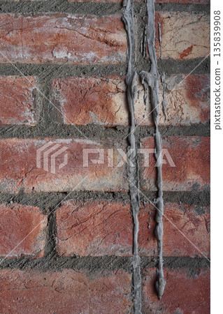 Brickwork made of hand-molded red brick and hand-mixed cement mortar. Gray cement streaks, reminiscent of melted candle wax, drip down the brick wall. Close-up. Abstract punk rock-style background. Brickwork made of hand-molded red brick and hand-mixed cement mortar. Gray cement streaks, reminiscent of melted candle wax, drip down the brick wall. Close-up. Abstract punk rock-style background. 135839908