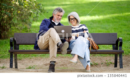 Senior couple using laptop sitting on park bench Senior couple using laptop sitting on park bench 135840441