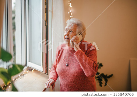 Old woman standing by window, making phone call. Old woman standing by window, making phone call. 135840706