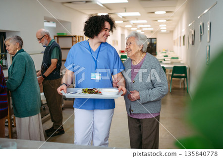 Senior woman having lunch with supportive caregiver in community center cafeteria. Senior woman having lunch with supportive caregiver in community center cafeteria. 135840778