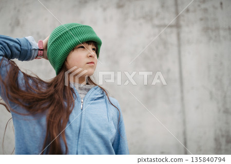 Outdoor portrait of cute young girl with beanie hat. Girl with long hair standing on playground in the city. 135840794