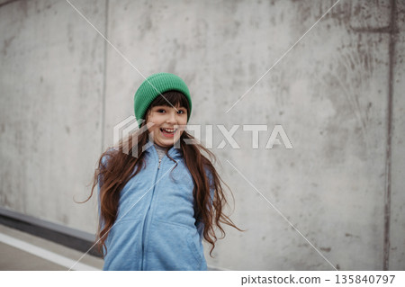 Outdoor portrait of cute young girl with beanie hat. Girl with long hair standing on playground in the city. 135840797