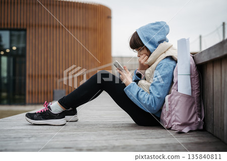 Girl sitting in front of school building, scrolling on phone. Online bullying or cyberbullying for young schoolgirl. Girl sitting in front of school building, scrolling on phone. Online bullying or cyberbullying for young schoolgirl. 135840811