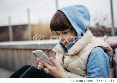 Girl sitting in front of school building, scrolling on phone. Online bullying or cyberbullying for young schoolgirl. Girl sitting in front of school building, scrolling on phone. Online bullying or cyberbullying for young schoolgirl. 135840813