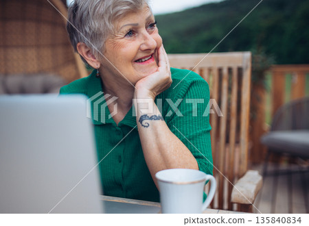 Mature woman sitting on balcony with laptop, working remotely. Mature woman sitting on balcony with laptop, working remotely. 135840834
