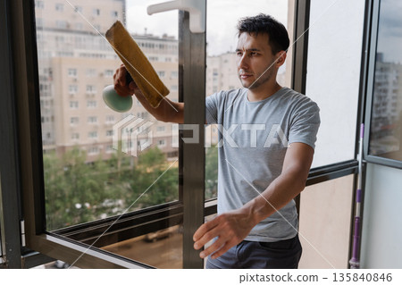 Young focused man wearing casual clothing cleaning large window in modern apartment, using squeegee to ensure spotless finish, with cityscape visible through glass. Concept of hygiene and cleanliness. 135840846