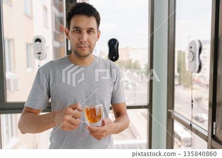 Portrait of handsome man looking at camera and holding cup of tea while robotic window cleaners work, showcasing convenience of modern smart home technology in contemporary apartment setting. 135840860