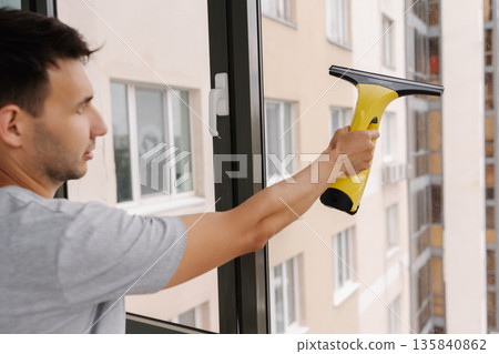 Back view of professional cleaning service worker washing windows in high-rise building using cordless electric window wiper ensuring streak-free and sparkling clean glass surfaces , close-up. 135840862