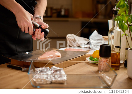 Woman seasoning raw chicken fillets on foil at kitchen table Woman seasoning raw chicken fillets on foil at kitchen table 135841264