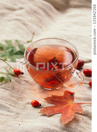 Rosehip tea in a glass cup on a linen tablecloth with autumn leaves and berries, cozy seasonal beverage. Rosehip tea in a glass cup on a linen tablecloth with autumn leaves and berries, cozy seasonal beverage. 135842108
