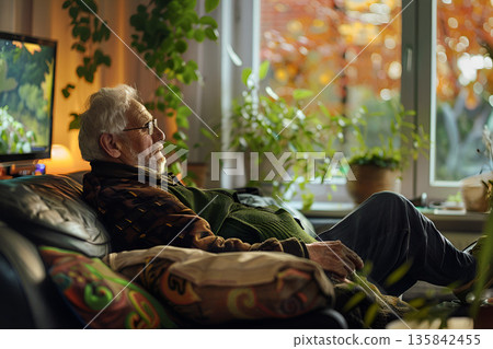 An elderly man is seated on a sofa near a window, looking outside 135842455