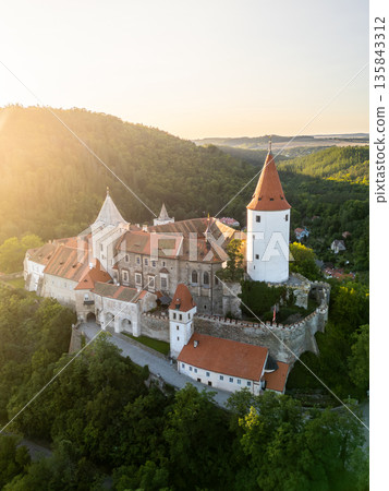 Krivoklat Castle showcases its medieval architecture in Central Bohemia during a stunning sunset. The aerial view highlights the castle's towers surrounded by lush green forests. 135843312