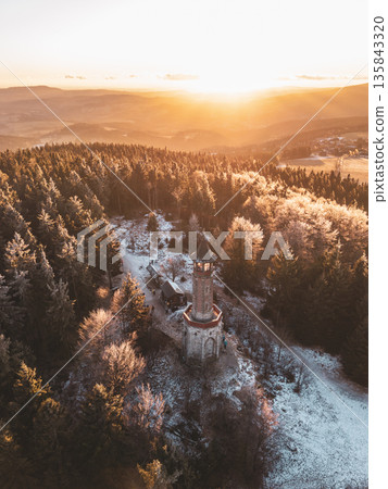 Stepanka lookout tower stands tall amidst the Jizera Mountains during winter. The sun sets behind the hills, casting light across snowy trees. The area feels quiet and still. Stepanka lookout tower stands tall amidst the Jizera Mountains during winter. The sun sets behind the hills, casting light across snowy trees. The area feels quiet and still. 135843320