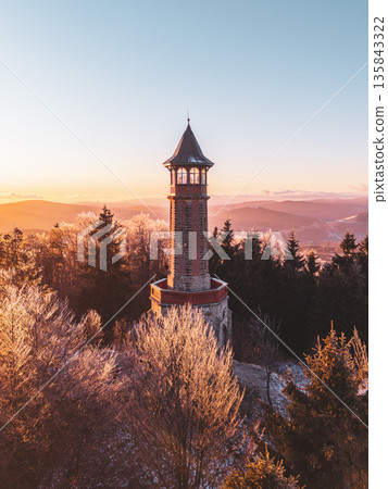 Stepanka lookout tower stands tall in the Jizera Mountains during winter. Frost covers the trees as the sun sets, casting warm light on the landscape. A peaceful scene invites exploration. 135843322